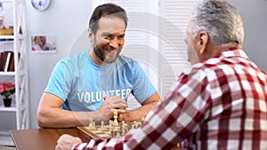 Smiling middle aged male volunteer playing chess with senior man in nursing home