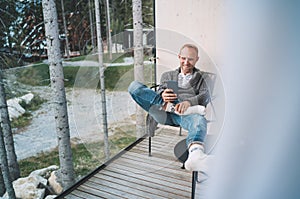 Smiling middle-aged man dressed open cardigan, jeans, and warm socks sitting on forest house balcony and using modern smartphone.