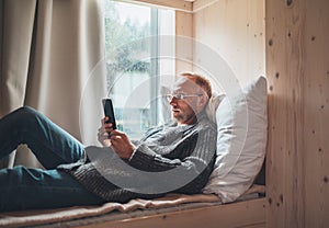 Smiling middle-aged man dressed warm knitted cardigan, jeans lying on cozy bed next to window and using modern smartphone.