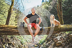 Smiling middle-aged man sitting on the fallen tree log over the mountain forest stream with his beagle dog while he waiting for