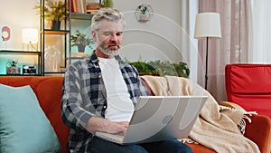 Smiling middle-aged mature man typing on laptop browsing internet using apps sitting at home sofa