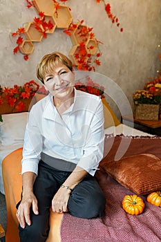 Smiling middle aged mature woman looking at camera, sitting at home on the bed with autumn decorations and pumpkins, thanksgiving