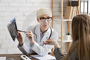 Smiling middle aged medical woman in glasses and white coat points to x-ray and communicates with patient