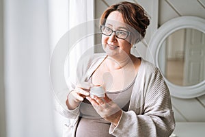 Smiling middle aged plus size woman with day cream near window at home