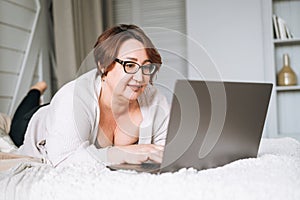 Smiling middle aged plus size woman lying on bed, working on laptop from home