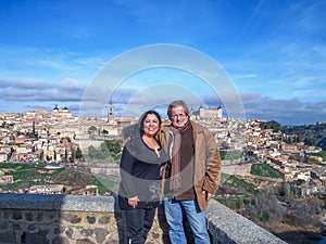 Smiling middle-aged tourist couple standing at viewpoint against blue sky, Toledo cityscape in background