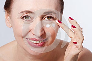 Smiling Middle aged woman applying cosmetic cream by finger on her face, white background. Macro and selective focus. Menopause an