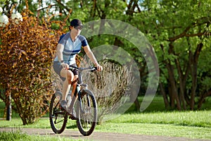 Smiling middle aged woman in cap with glasses cycling in summer park, cycling cardio training, city walks with bike