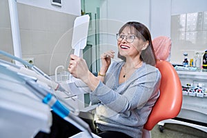 Smiling middle aged woman in dental chair with mirror looking at her teeth
