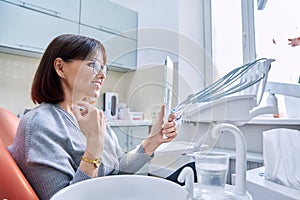 Smiling middle aged woman in dental chair with mirror looking at her teeth