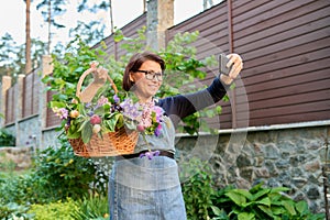 Smiling middle aged woman in garden with basket of fresh cut flowers with smartphone.