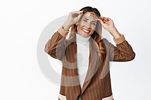 Smiling middle aged woman take on eyewear and looking happy, trying new glasses, standing in brown suit against white