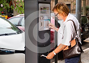 Smiling middle aged woman using parking machine to pay for car parking on summer city street