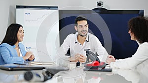 Smiling Middle Eastern young man listening to excited Asian woman talking and gesturing sitting at conference table with