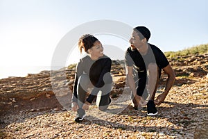 Smiling millennial african american lady and man in sportswear tying shoelaces on shoes on ocean beach