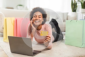 Smiling millennial african american woman with many packages shows credit card uses laptop, lies on floor