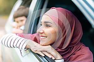 Smiling millennial arab female in hijab and boy look out the open window and enjoy journey in car