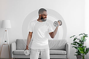 Smiling millennial bearded african american guy in white clothes lifting dumbbells in living room