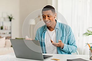 Smiling millennial black guy in jeans with wireless headphones has video call in room office interior