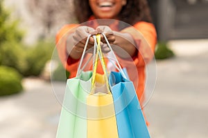 Smiling millennial black woman in casual shows many of bags with order, enjoys great shopping in city, cropped