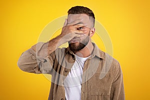 Smiling millennial caucasian guy with beard in casual covers face with hand, spies with eye