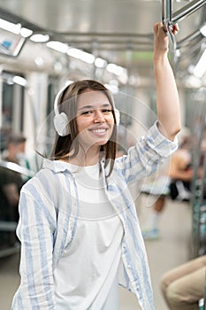Smiling millennial cute girl passenger listening to music with wireless earphones in subway train.