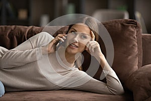 Smiling millennial female lying on comfy sofa talking on telephone