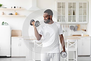 Smiling millennial handsome bearded african american male athlete in white clothes lifts dumbbells in minimalist kitchen