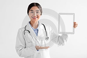 Smiling millennial indian female doctor in coat shows on tablet with blank screen isolated on white background