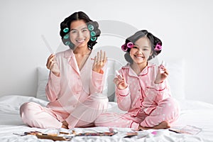 Smiling millennial japanese female and teen girl in pajamas and curlers do manicure on bed in bedroom