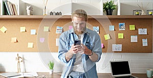 Smiling millennial man standing at home office workspace using cell phone.