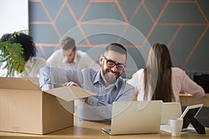 Smiling millennial new male employee unpacking box at office wor