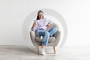 Smiling millennial woman chatting online on smartphone, sitting in comfy armchair on white studio background