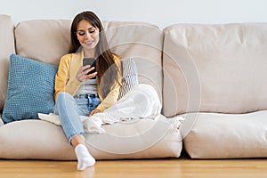 Smiling millennial woman sit on couch at home, using phone, texting chatting with friend. Happy girl resting on sofa hold