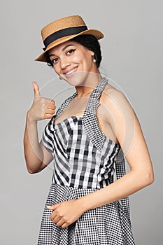 Smiling mixed race woman in chequered summer dress and canotier straw hat standing gesturing thumb up