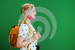 Smiling modern girl with backpack and mask isolated on green