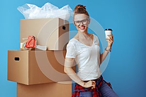 Smiling modern woman near cardboard box with coffee cup on blue