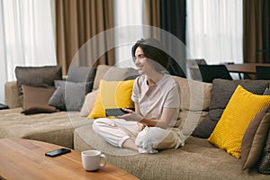Smiling modern young girl holding TV remote control watching series or movie, sitting on comfortable sofa at home