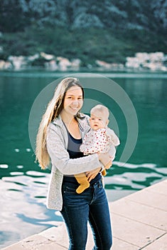 Smiling mom with a baby in her arms stands on the pier