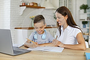 Smiling mom is helping her son doing homework studying online using laptop in home kitchen.