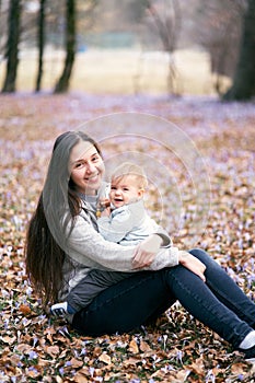 Smiling mom holding a laughing baby in her arms sitting on the grass among small wildflowers in the park