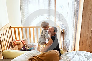 Smiling mom lies on the bed near the standing baby on the background of a crib and toys