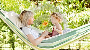 Smiling mom and little girl daughter child blue eyes with blond curly hair, together lying on the hammock in the green home garden