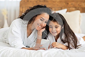 Smiling mom and little girl laying on bed, using smartphone