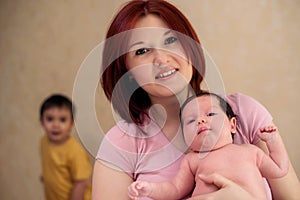 Smiling mom posing with infant child. Older kid is standing left behind, out of focus. Parenting issues when new baby arrives