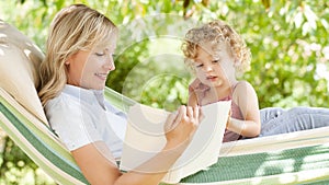 Smiling mom reading a story from the book to the little girl daughter child blue eyes with blond curly hair, together lying on the