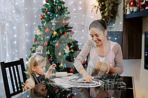 Smiling mom rubbing flour on table holding dough in hand while sitting next to little girl