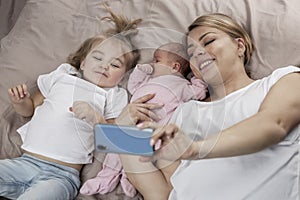 Smiling mom with small children lies on the bed and takes a selfie on the phone. Love and tenderness