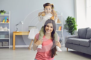 Smiling mother carrying cute little daughter on her shoulders at home. Parent and child with toy playing indoors