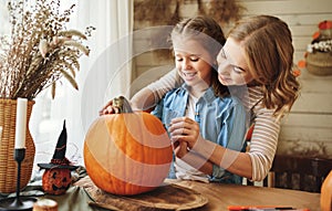 Smiling mother and child girl drawing scary faces on Halloween pumpkins while sitting in cozy kitchen